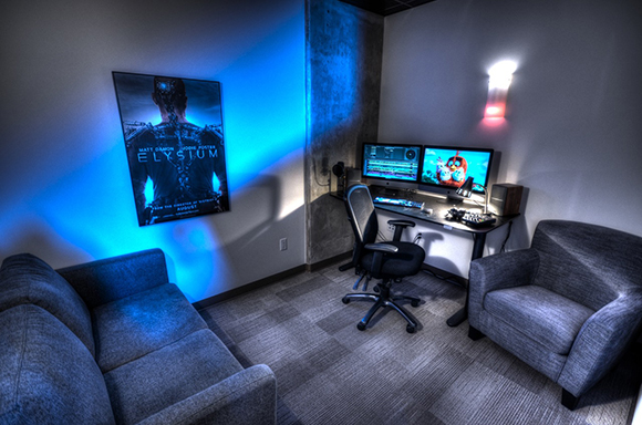 A dark room featuring a desk with dual monitors, an office chair, a sofa, and an armchair under blue lighting.