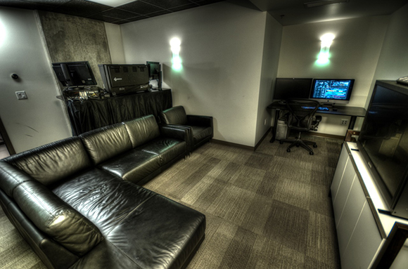 A lounge area with a black leather sectional sofa, a desk with computer monitors, and a large television.