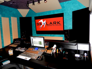 A recording studio desk with a mixing console, laptop, and a screen displaying the Skylark Sound Studios logo.