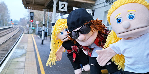 Three puppets with blonde and brown hair posing on a wet train platform next to tracks.
