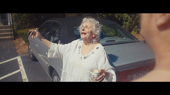 An elderly woman in a white shirt smiles and gestures while holding a mug next to a grey car.