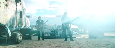 Two musicians play electric guitars in front of a drum kit near a large, weathered vintage airplane.