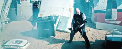 A bald man in black clothing plays an electric guitar amidst airplane wreckage in a sandy desert.