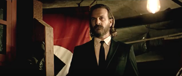 A bearded man in a black suit and tie stands before a red and white flag in a dimly lit room.
