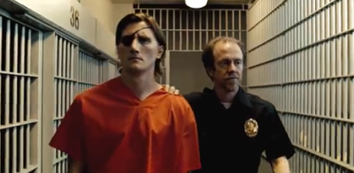 A man in an orange jumpsuit and eye patch is escorted by a guard through a prison cell block.