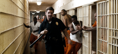 A prison guard in a black uniform runs through a cell block as inmates react from their cells.