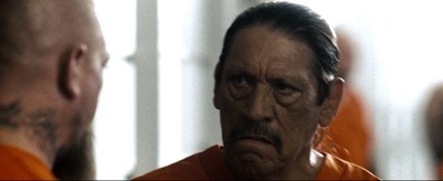A man with dark hair and a mustache in an orange shirt looks intensely at another person.