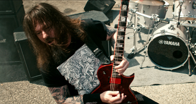 A long-haired musician playing a red electric guitar with a drum kit in the background.
