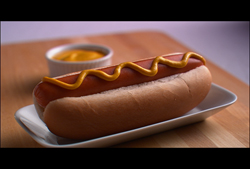 A hot dog in a bun topped with a wavy line of yellow mustard, served on a white plate next to a small bowl of mustard.