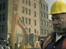 A construction worker wearing a yellow hard hat and safety vest looks forward near an excavator and city buildings.