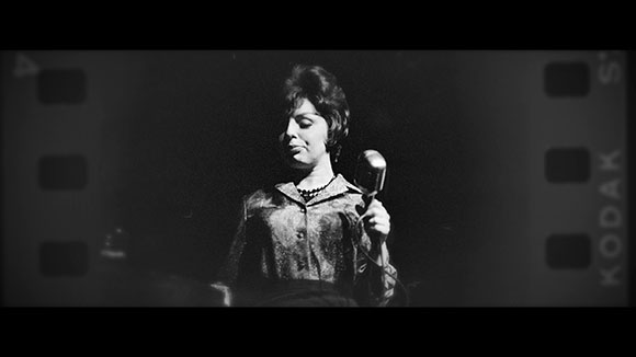 A woman with dark hair sings into a vintage microphone against a dark background.