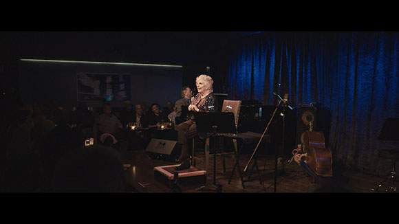 A woman with white hair sings into a microphone on a dark stage before an audience.