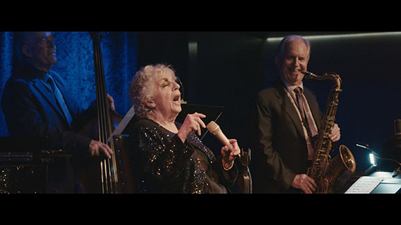 An elderly woman sings into a microphone while a man plays saxophone and another plays double bass.
