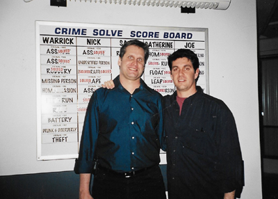 Two men smiling while standing in front of a large white crime solve score board.
