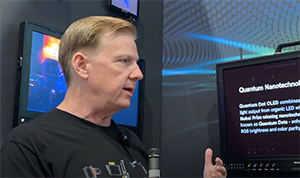 A man in a black t-shirt gestures while speaking next to a screen displaying text about Quantum Nanotechnology.