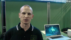 A man in a black polo shirt stands in front of a green screen next to a laptop displaying a graphic.