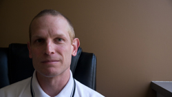 Close-up of a man with short hair and a light complexion, wearing a white collared shirt and a lanyard.