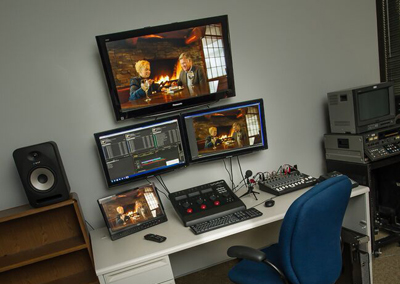 A video editing workstation with four monitors displaying footage, a keyboard, a control surface, a small audio mixer, and a