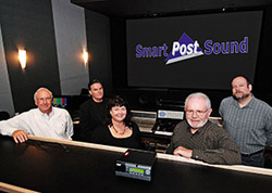 Five people smiling behind a mixing console in a dark sound studio with a large screen.