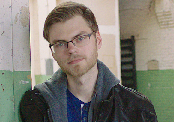 A man with light brown hair, glasses, and a beard wearing a black leather jacket over a grey hoodie.