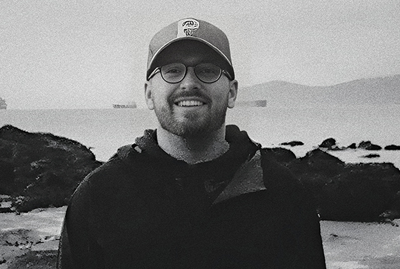 A smiling man with glasses and a baseball cap stands in front of a coastal landscape.