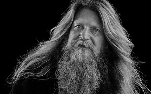 Black and white close-up portrait of a man with long, wavy hair and a thick, textured beard.
