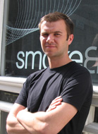 A man with short brown hair wearing a black t-shirt stands with his arms crossed in front of a glass window.