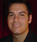 A smiling man with dark hair wearing a black collared shirt against a dark red background.