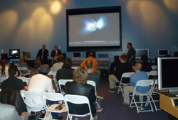 Audience watches a presentation on a large screen in a room with computers.