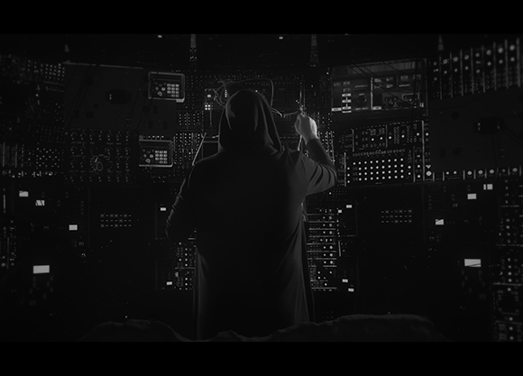 Black and white shot of a person in a dark hood facing a large wall of illuminated electronic control panels.