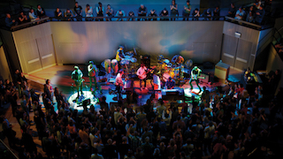 A musical band performs on a brightly lit stage before a large seated and standing audience in a dark venue.