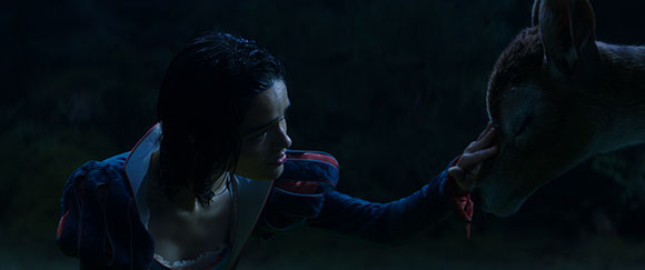 A woman with dark hair reaches out to touch the face of a deer in a dark setting.