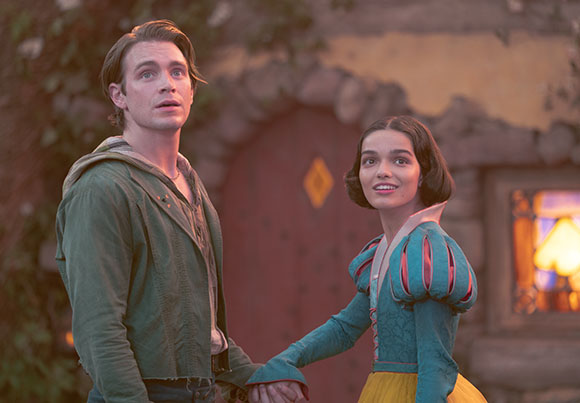 A man in a green tunic and a woman in a teal and yellow dress hold hands while looking up in awe.
