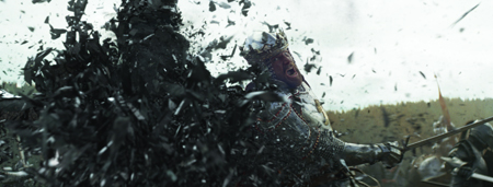 A person in silver armor and a crown shatters into many small black fragments against a cloudy sky.