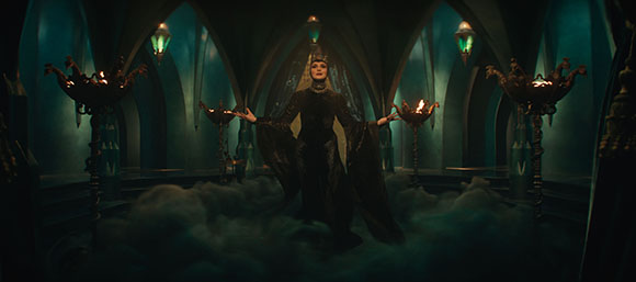 A woman in a dark, flowing gown stands amidst fog in a dark, arched hall with glowing lanterns.
