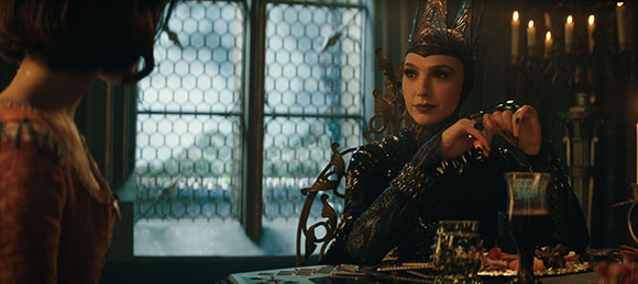 A woman in a dark crown and ornate black clothing sits at a table in a room with leaded glass windows.