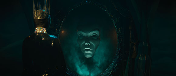 A person wearing a crown looks at a pale mask inside a glowing circular frame in a dark setting.