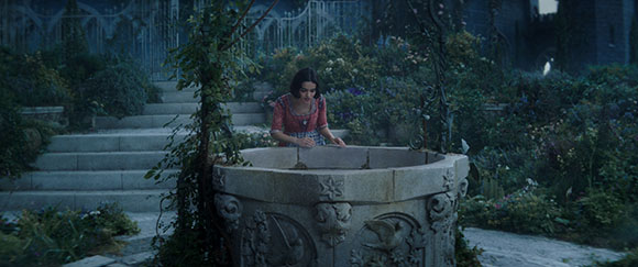 A woman in a red dress leans over an ornate stone well in a lush, misty garden.