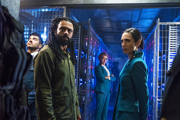 Four people stand in a blue-lit hallway lined with metal storage cages.
