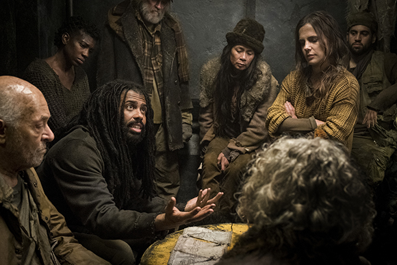 A group of people in worn clothing sit in a dark room, listening to a man gesturing with his hands.