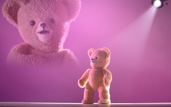 A small teddy bear stands on a stage under a spotlight with a large teddy bear projection in the background.