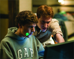 Two young men lean in closely to look at a glowing computer monitor in a dimly lit room.