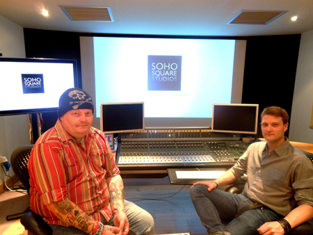 Two men sit in a professional recording studio with a large mixing console and a screen displaying a logo.