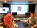 Two men sit in a room facing large projection screens and monitors.