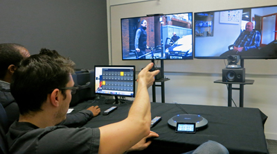 A person in a dark shirt points toward two large monitors displaying video footage in a studio.