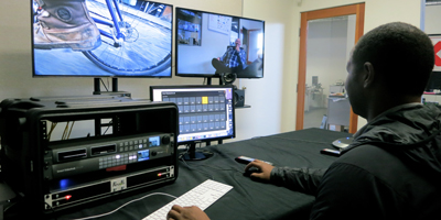 A person sits at a desk using a keyboard and mouse, facing three computer monitors and a rack of video equipment.