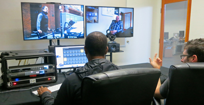 A person sits in front of a large monitor displaying a video of a man in a dark shirt.