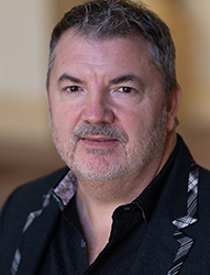 A middle-aged man with short gray hair and a salt-and-pepper beard wearing a black patterned jacket.