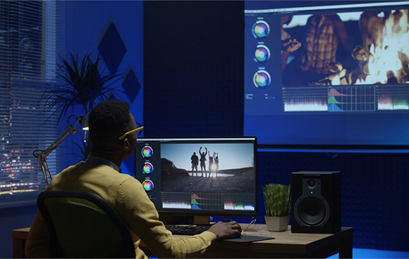 A person sits at a desk with a computer monitor and large screen displaying video editing software.