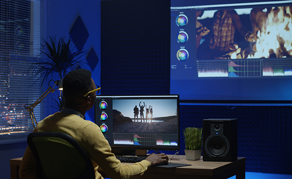 A person sits at a desk with a computer monitor and large screen displaying video editing software.